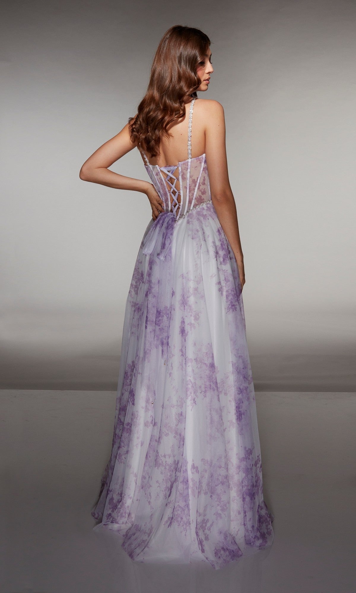 A woman faces away from the camera wearing the Alyce 61994 long prom dress, a sleeveless white gown with lavender floral patterns and a lace-up corset bodice.
